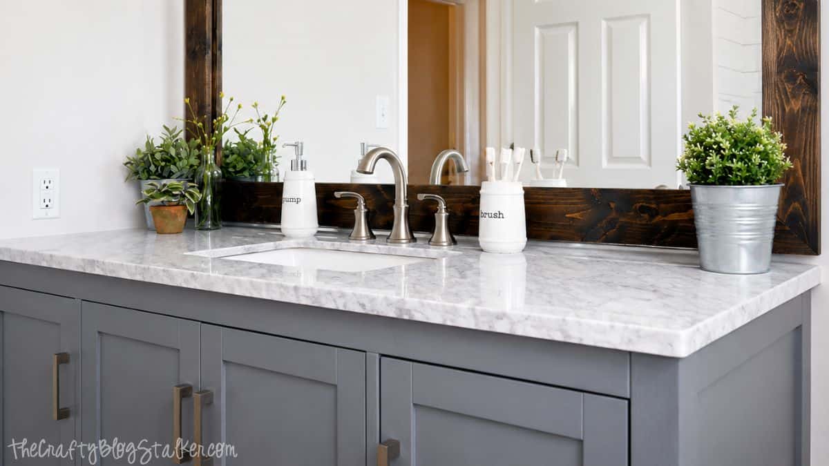 Gray bathroom vanity with marble-look countertop, brushed nickel faucet, wood-framed mirror, and modern bathroom decor.