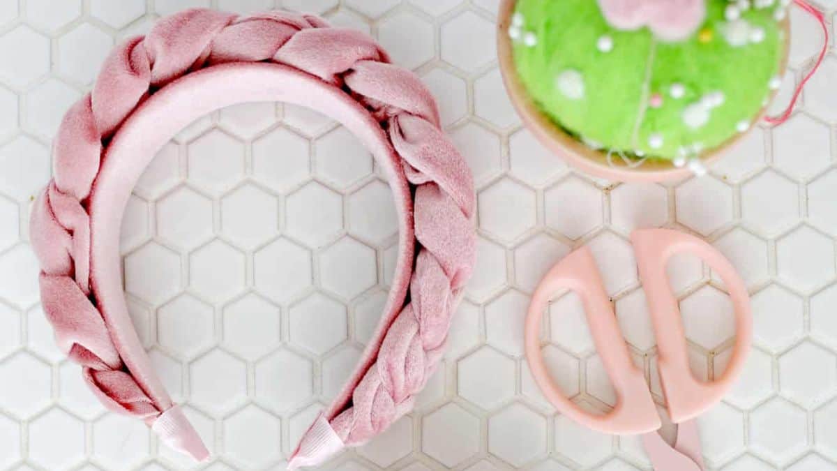 Pink braided velvet headband displayed beside pink scissors and a pincushion on a white hexagon tile background.