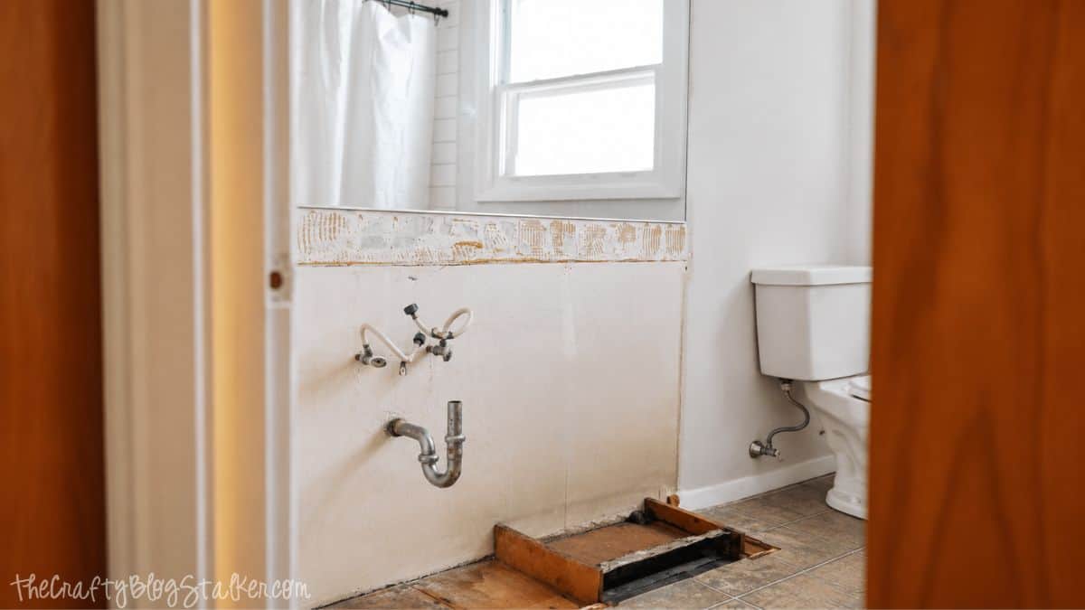 Bathroom wall with exposed plumbing and unfinished floor after removing an old vanity during a bathroom remodel.