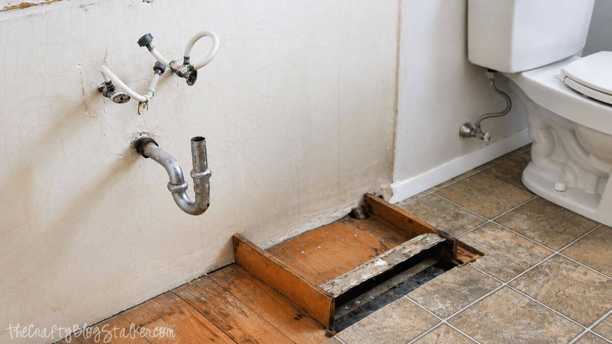 Bathroom with old vanity removed, showing exposed plumbing, damaged wall, and unfinished floor before installing a new vanity.