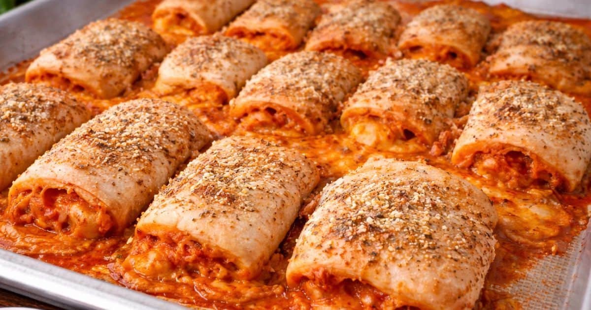 Baked pizza roll ups on a sheet pan, filled with cheese and sauce, topped with seasoning and melted cheese spilling out of the edges