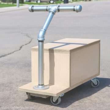 Homemade wooden ride-on toy with caster wheels and a metal pipe handlebar, sitting on pavement and built for kids to scoot and play.