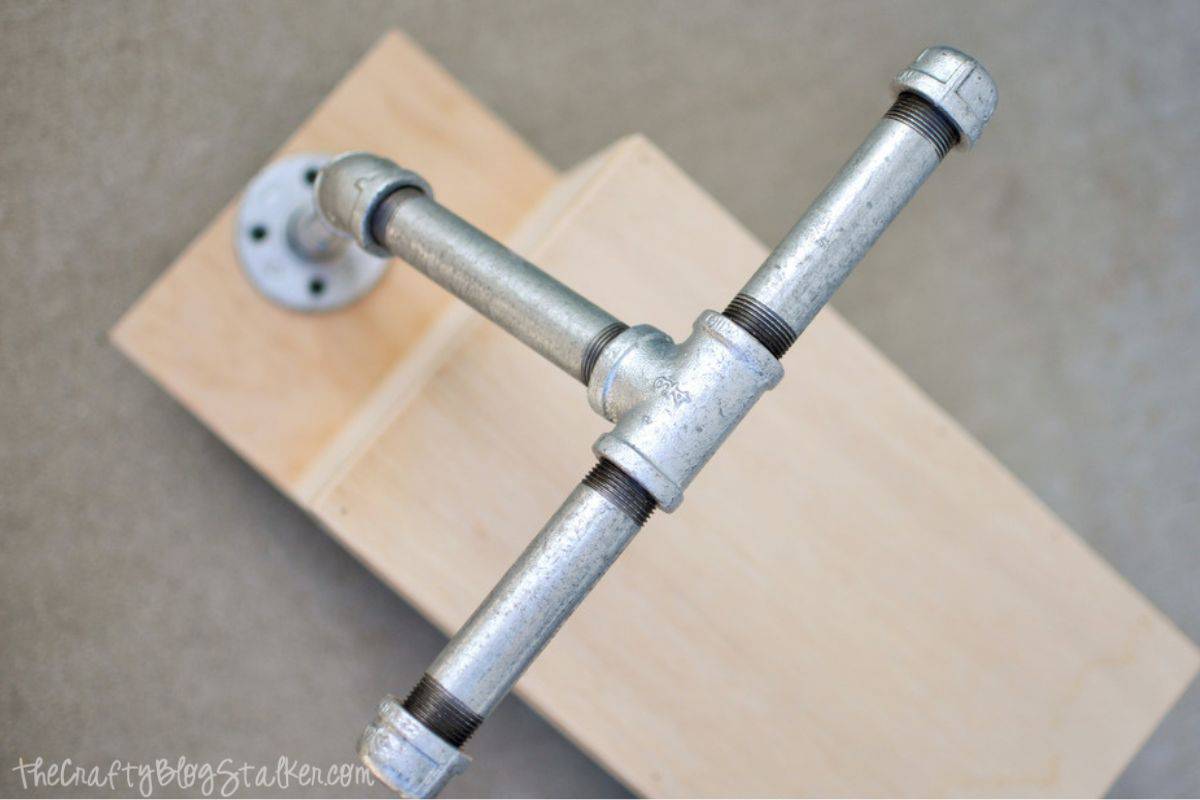 Close-up view of metal pipe handlebars attached to a wooden ride-on toy, showing the tee fitting, elbows, and flange mount.