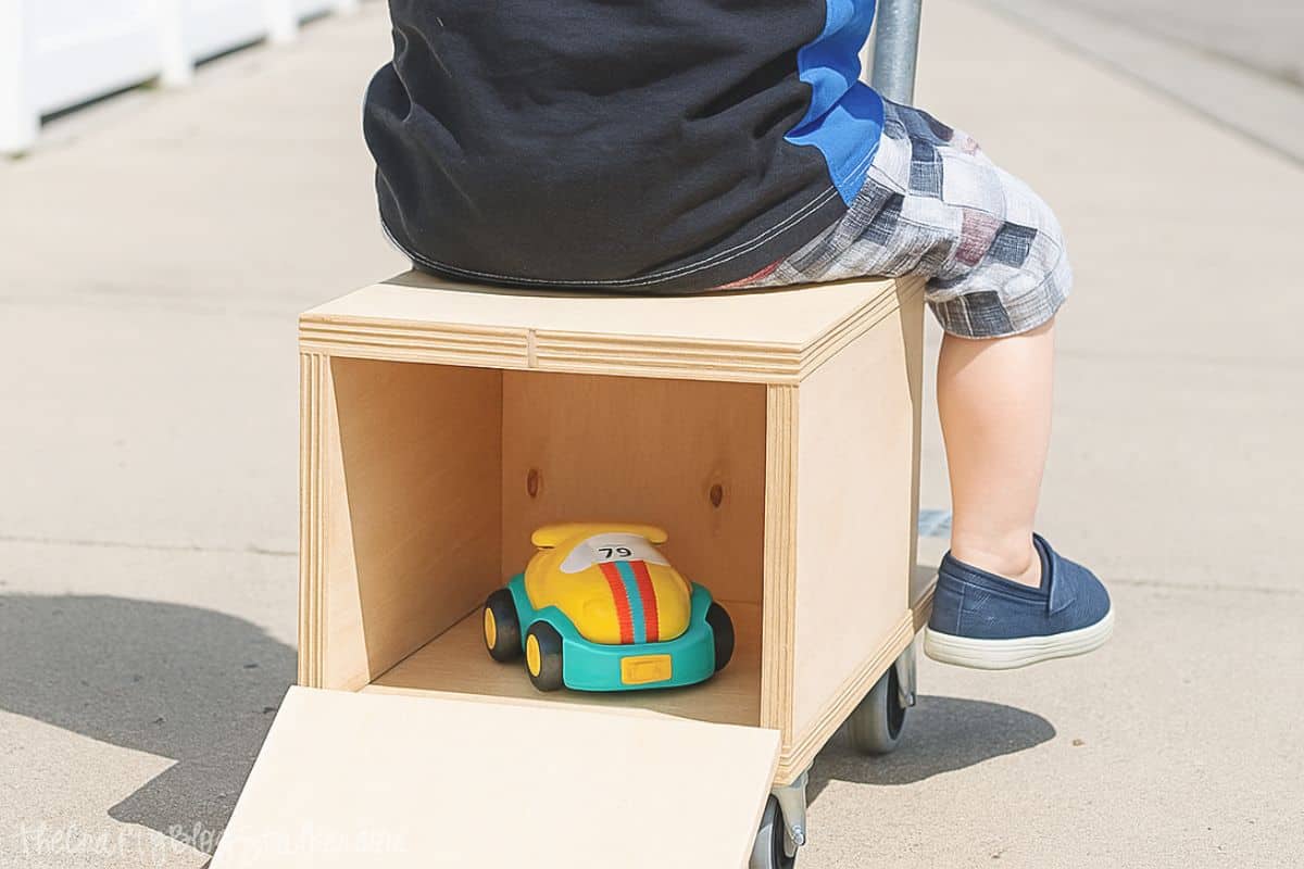 Child sits on a wooden ride-on toy with an open storage compartment in the back, revealing a colorful toy car inside.