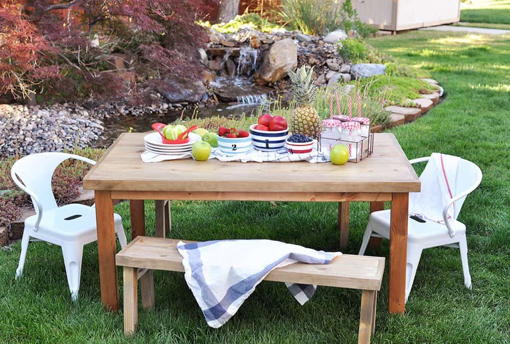Outdoor picnic setup with a wooden table and bench, white chairs, and a colorful fruit display near a waterfall in a landscaped backyard.
