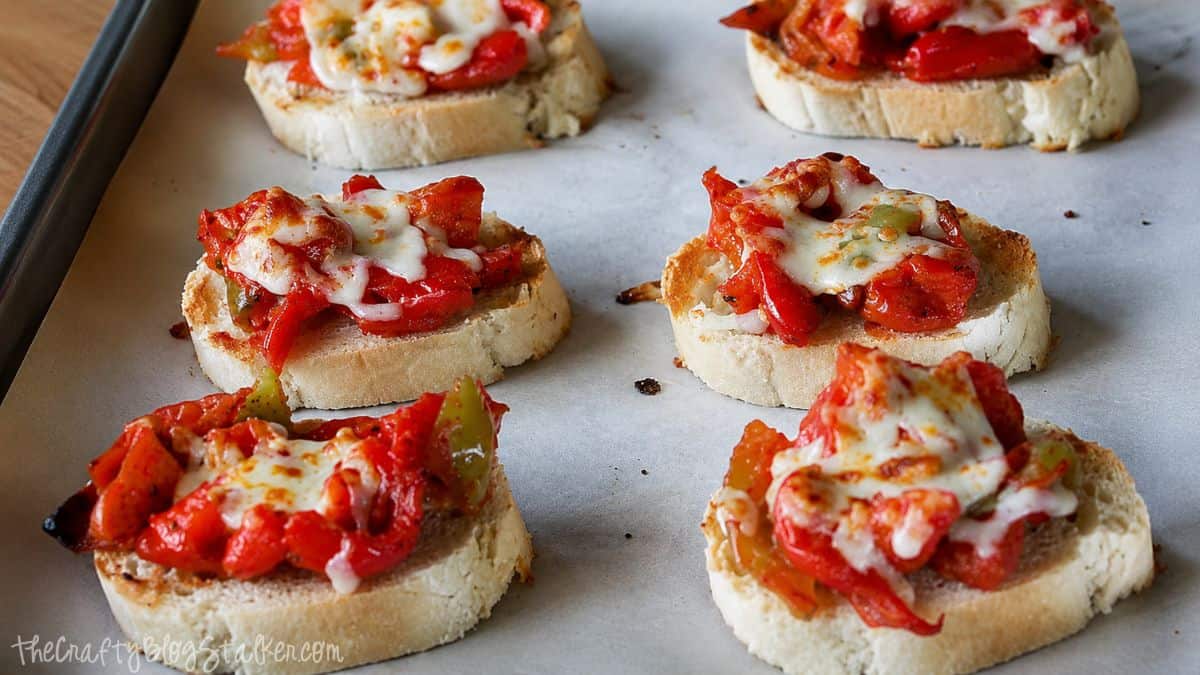 Close-up of sausage and pepper bruschetta topped with melted cheese on toasted bread slices on a baking sheet.