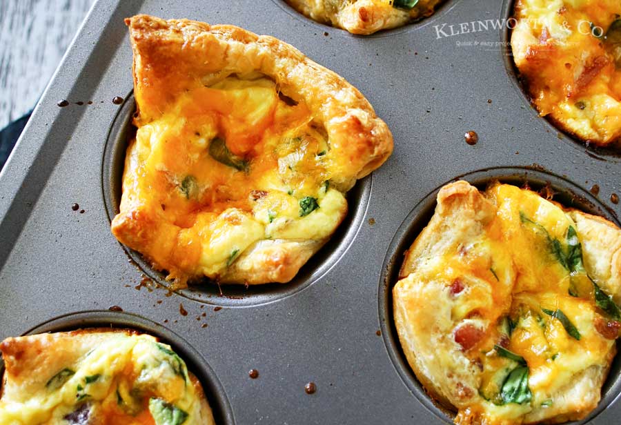 Mini puff pastry quiche cups baked in a muffin tin, filled with eggs, cheese, spinach, and bacon.