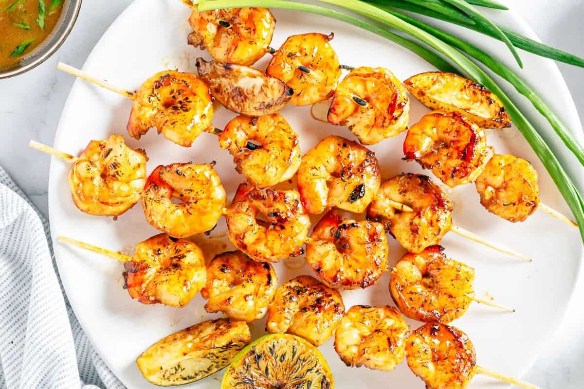 Grilled shrimp skewers with a golden Asian marinade, served on a white plate with charred lemon wedges and green onions as garnish.