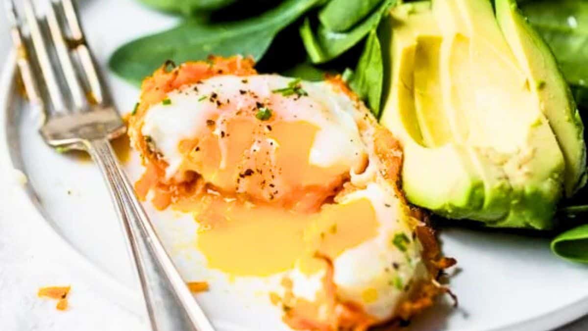 A plate with a baked sweet potato egg cup cut open to reveal a runny yolk, served alongside sliced avocado and fresh greens.