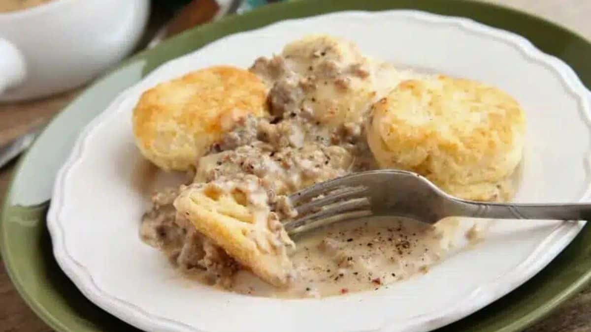 A plate of biscuits served with creamy sausage gravy, with a fork taking a bite from one of the biscuits.