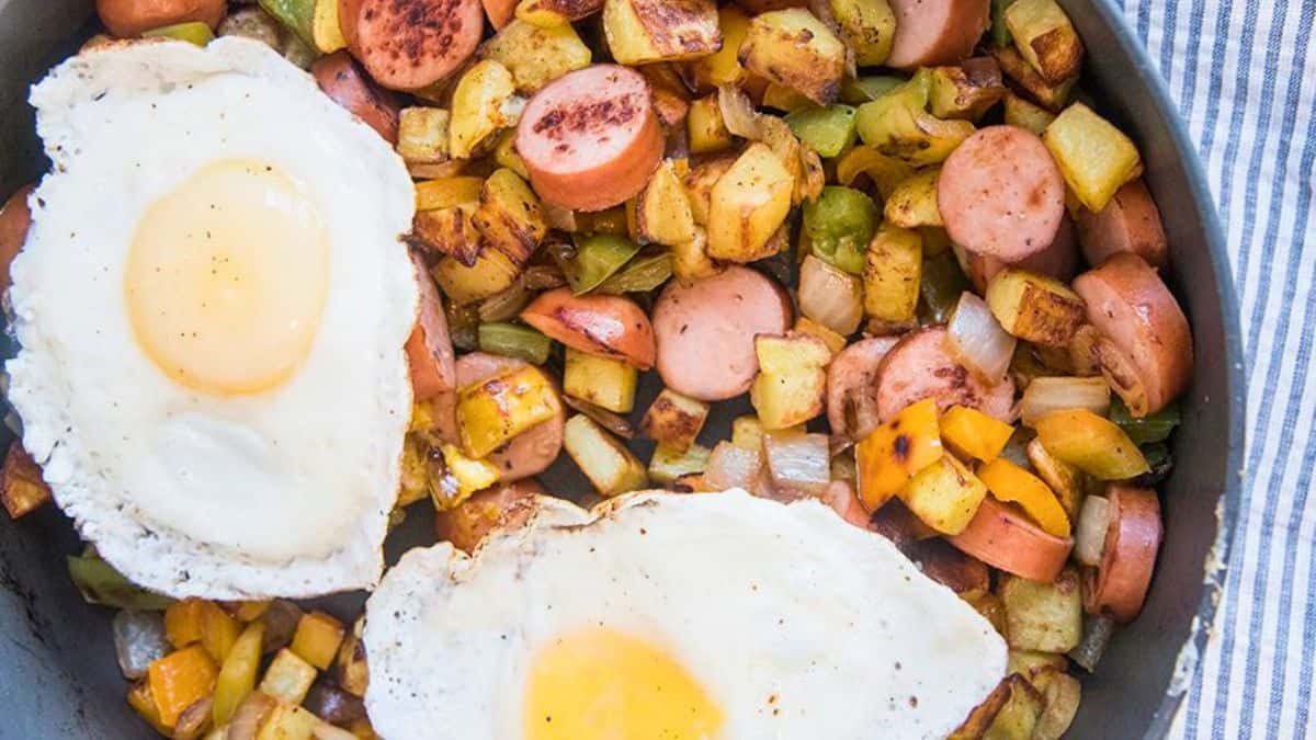 A skillet filled with breakfast hash made of fried potatoes, sausage slices, peppers, and onions, topped with two sunny-side-up eggs.