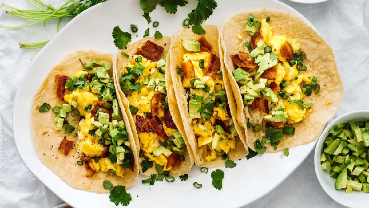 Four breakfast tacos filled with scrambled eggs, crispy bacon, avocado, and fresh herbs arranged on a white plate.