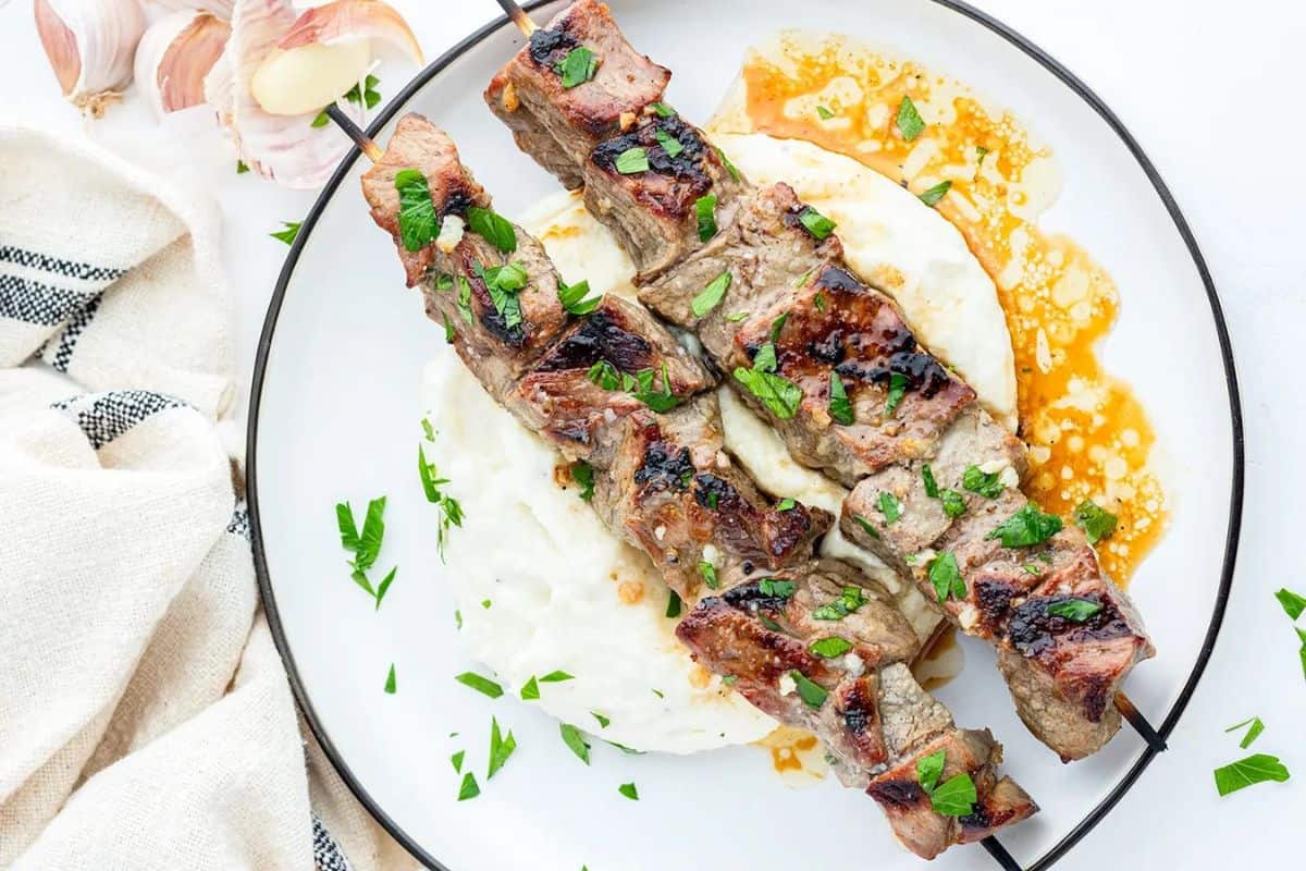 Grilled garlic sirloin skewers served over creamy mashed potatoes, garnished with chopped parsley and drizzled with a savory sauce on a white plate.