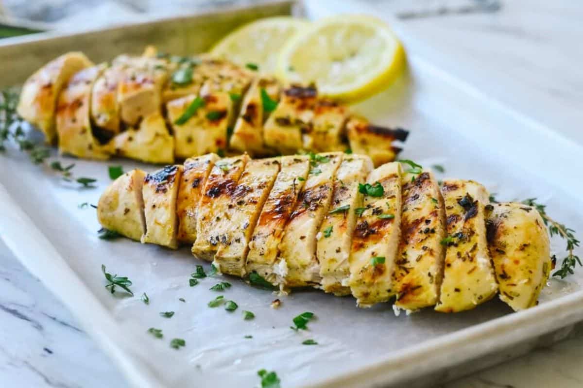 Sliced grilled lemon mustard chicken breast garnished with fresh herbs, served on a tray with lemon slices in the background.