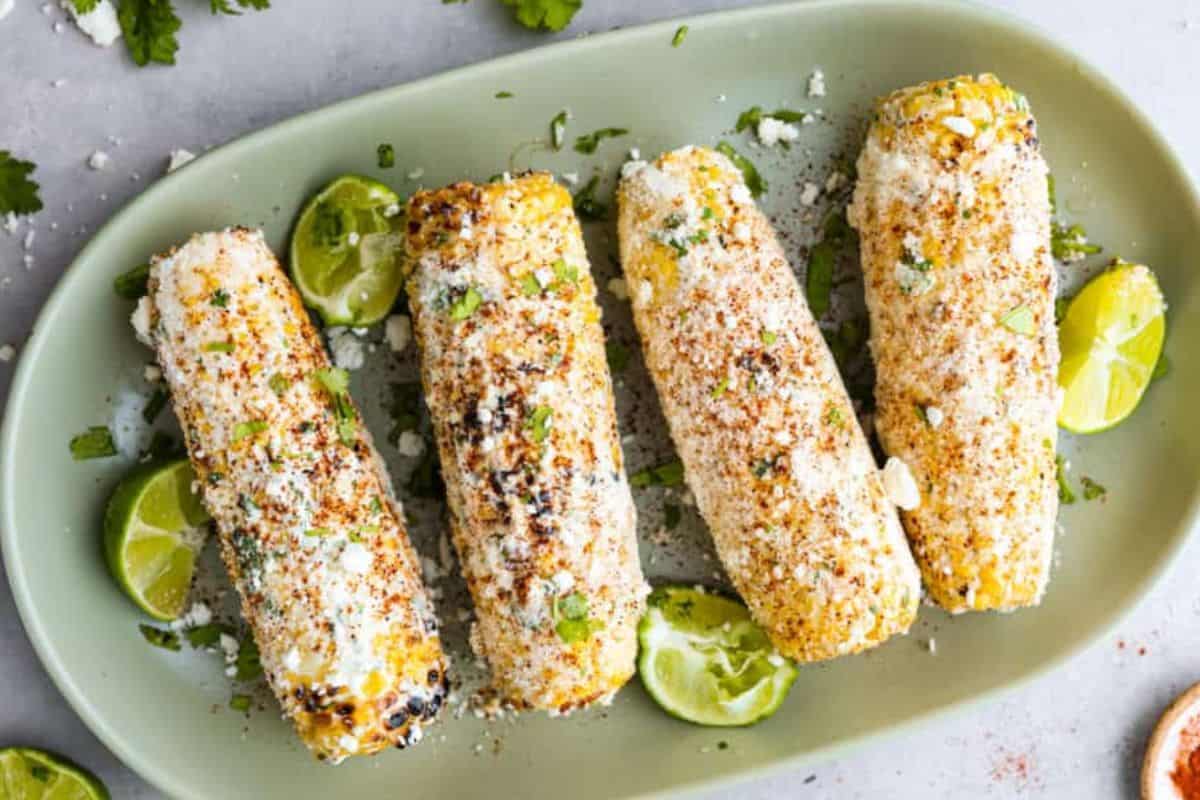 Four grilled Mexican street corn cobs topped with cotija cheese, chili powder, and chopped cilantro, served with lime wedges on a green plate.