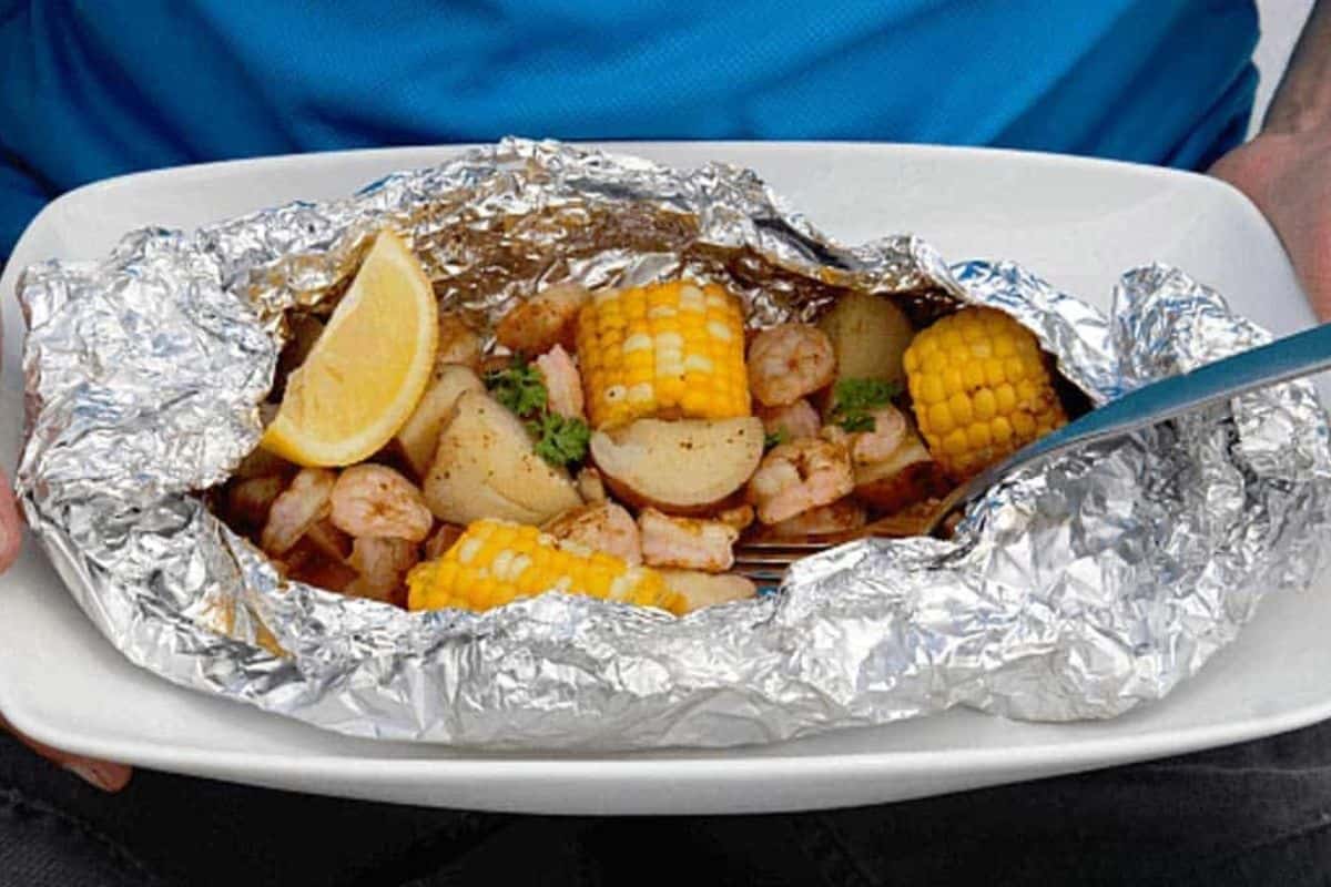Shrimp boil foil packet with shrimp, corn, potatoes, lemon wedge, and parsley served in foil on a white plate, held by someone in a blue shirt.