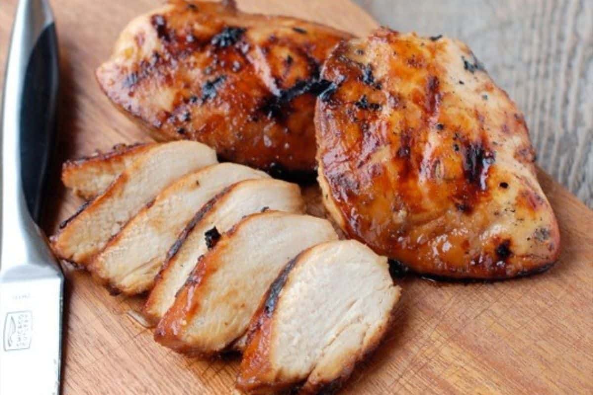 Sliced and whole grilled chicken breasts on a wooden cutting board, showcasing a juicy, golden-brown glaze with a knife placed nearby.