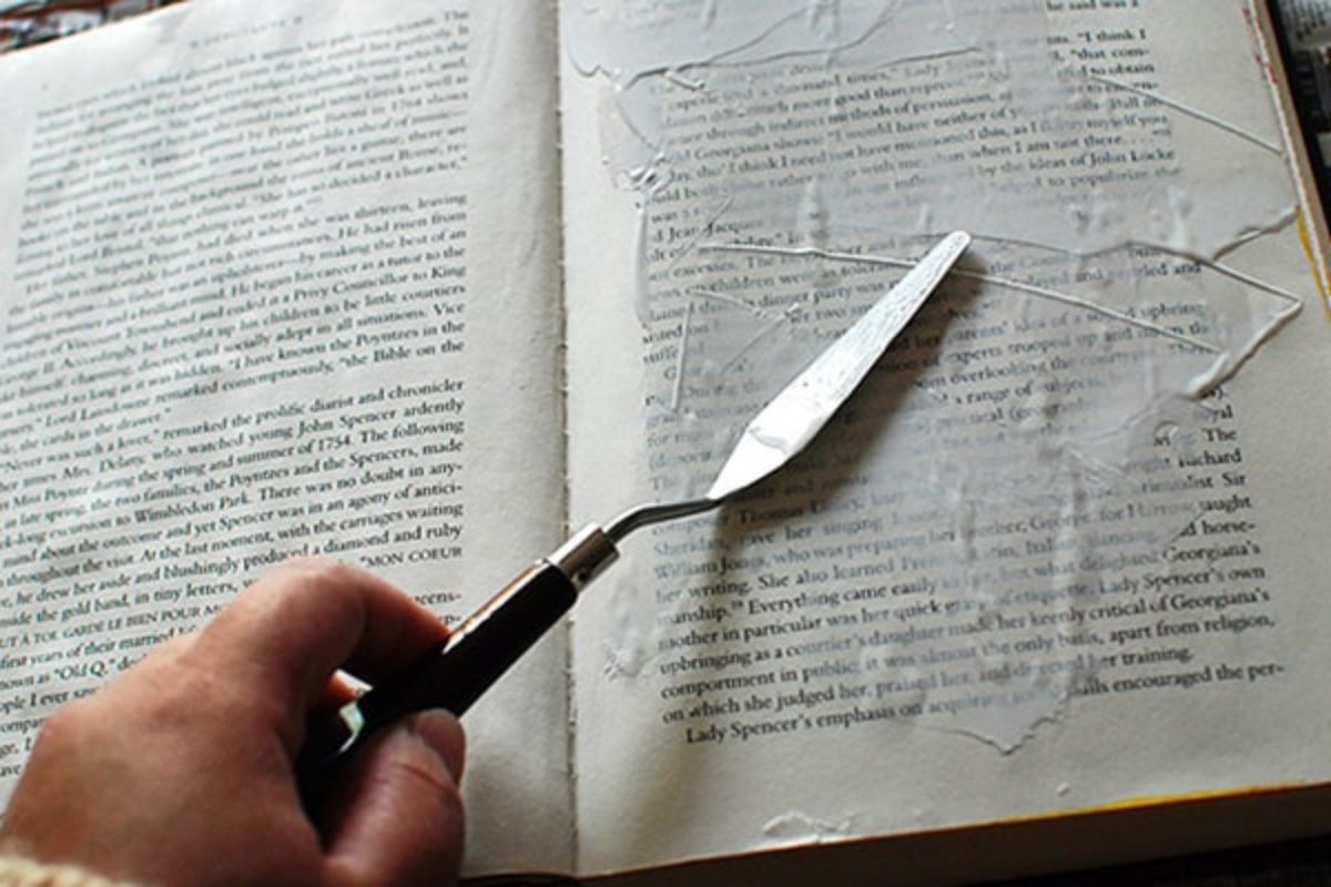 A hand uses a palette knife to spread white gesso over the text on a book page, preparing it for mixed media or art journaling.