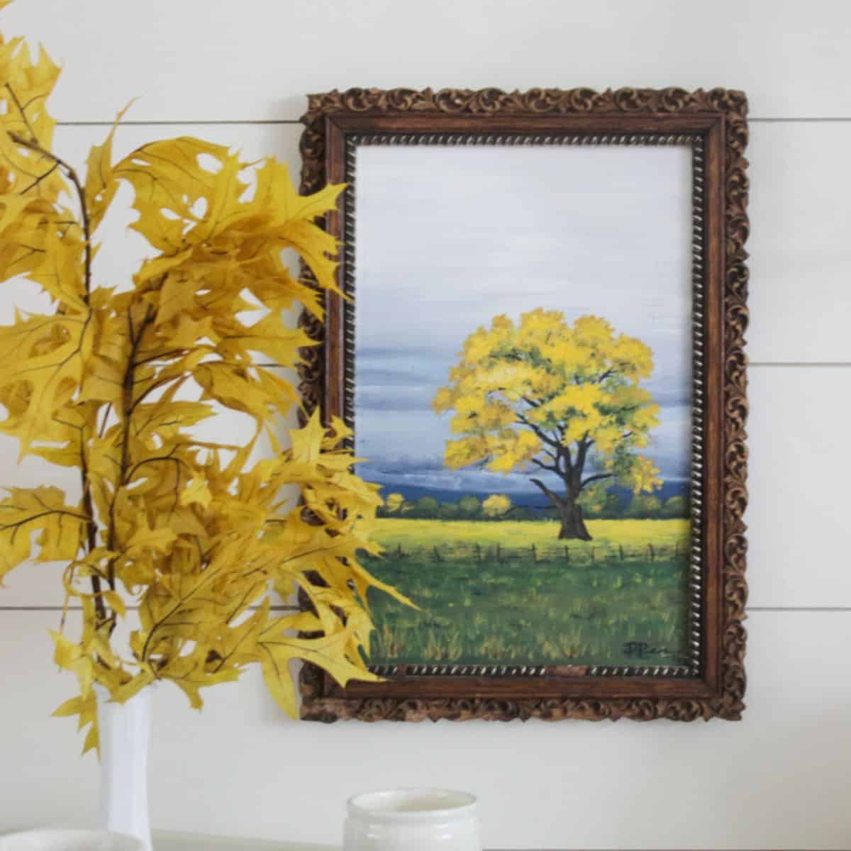 Framed autumn painting of a yellow-leaved tree in a green field, displayed beside a vase filled with golden fall branches.