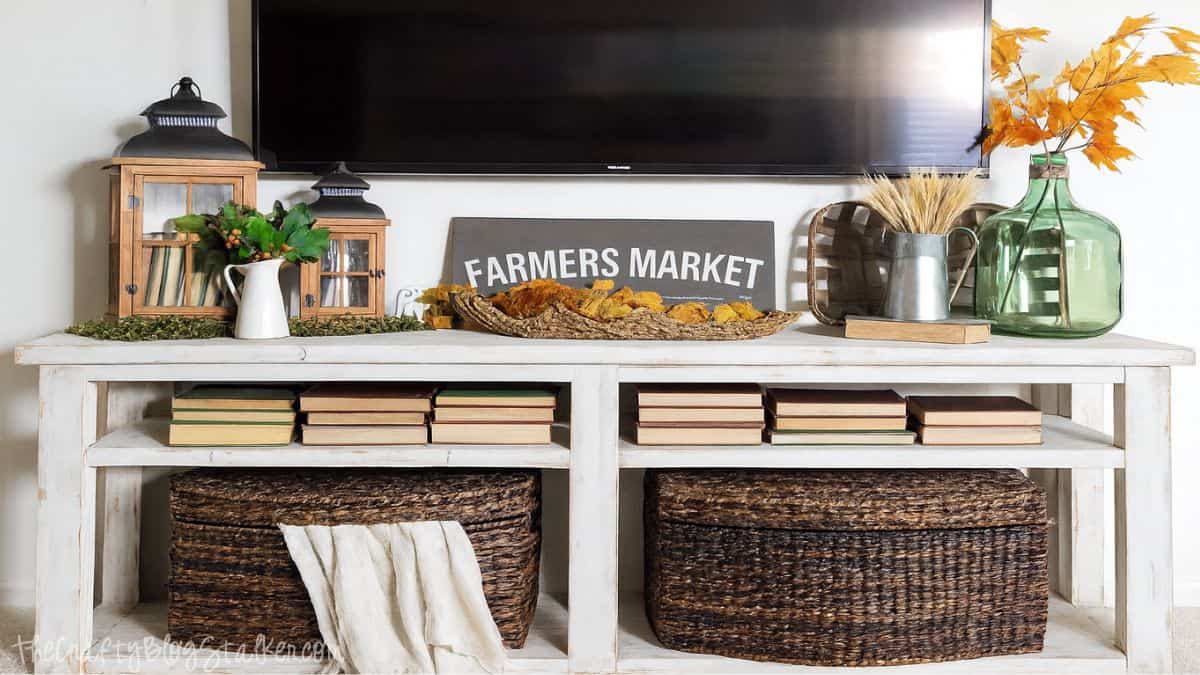 Rustic fall farmhouse console table decorated with lanterns, books, baskets, a farmers market sign, wheat, and autumn leaves.