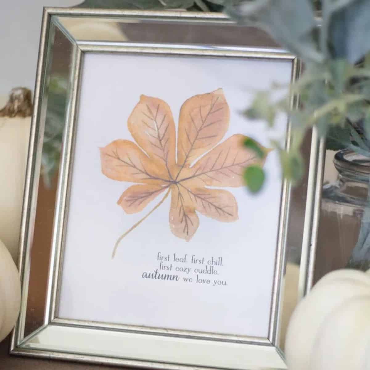 Framed fall printable featuring a watercolor autumn leaf and seasonal quote, displayed with white pumpkins and greenery.