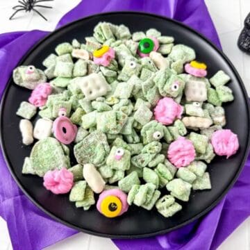 A black plate filled with green Halloween muddy buddies mixed with candy eyes, pink and yellow candies, and pretzels, set on a purple cloth.