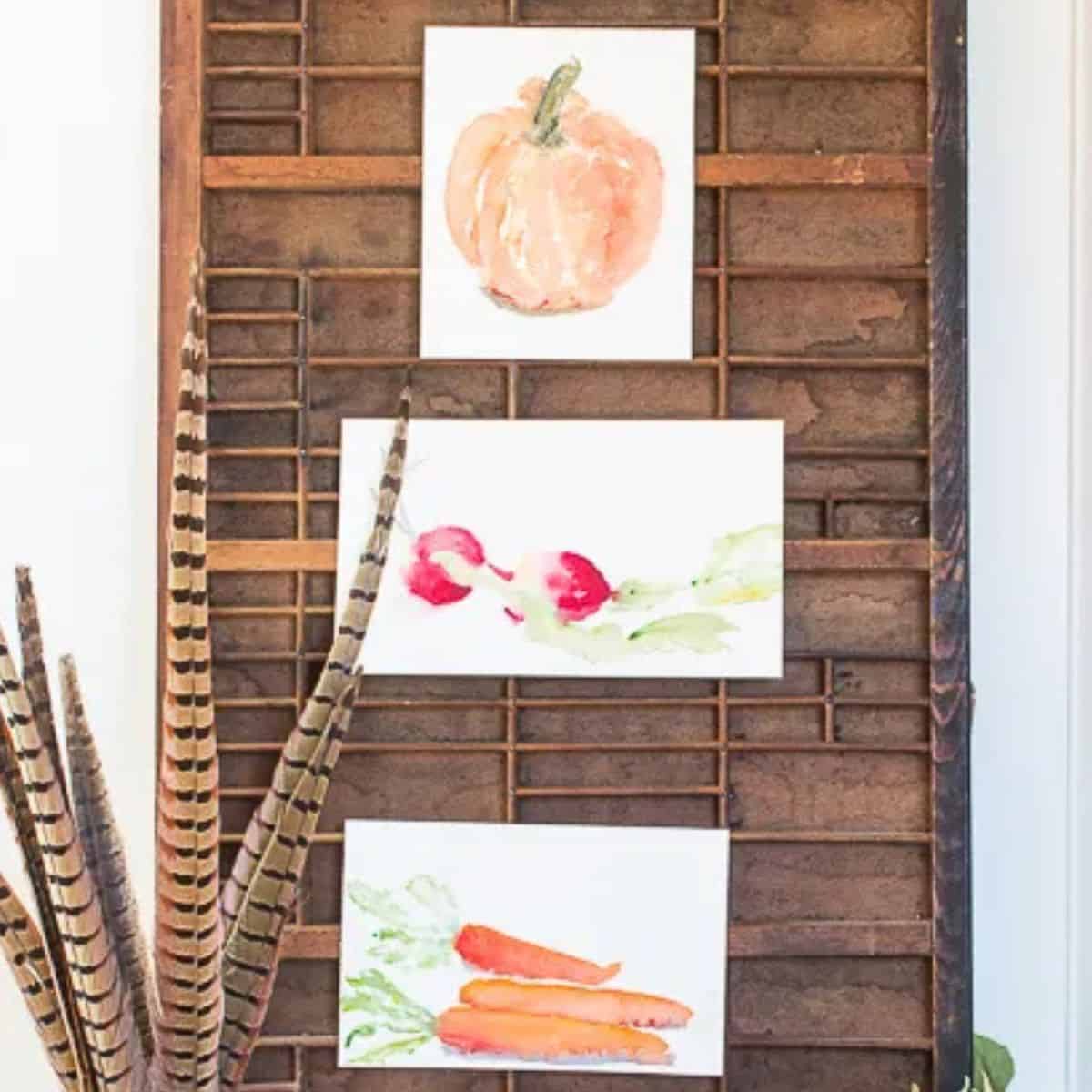 Three harvest watercolor prints featuring a pumpkin, radishes, and carrots displayed on a vintage wooden printer's tray.