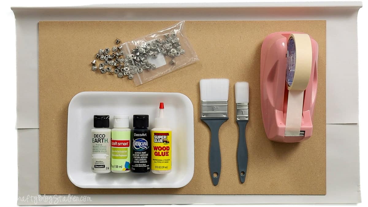 Craft supplies including MDF board, screws, paint bottles, wood glue, paintbrushes, masking tape, and a pink tape dispenser arranged for a DIY project.