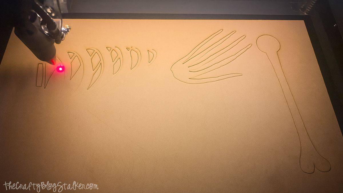 Laser cutting machine etching skeleton pieces onto a wood sheet, with visible outlines of bones including a hand and long bone shapes.