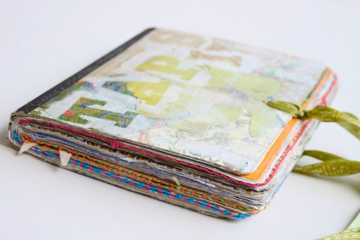 A handmade art journal with a colorful, textured cover and stitched spine, tied closed with a green ribbon, resting on a white surface.