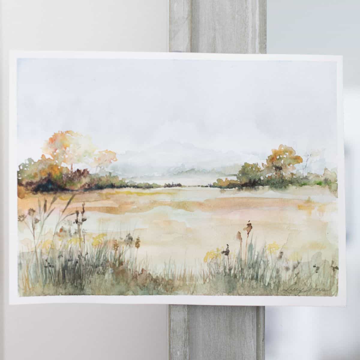 Watercolor fall landscape featuring a calm lake, golden autumn trees, soft distant hills, and grasses along the shoreline.