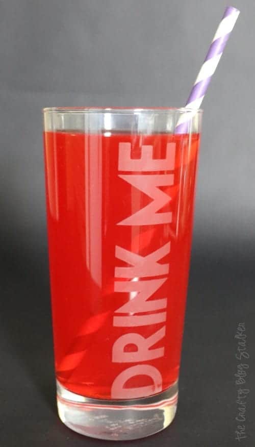 a "drink me" etched glass filled with a red juice and a purple striped straw
