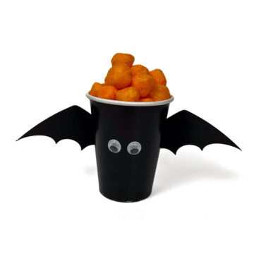 Black paper cup decorated as a bat with wings and googly eyes, filled with cheese balls for a fun DIY Halloween party treat.