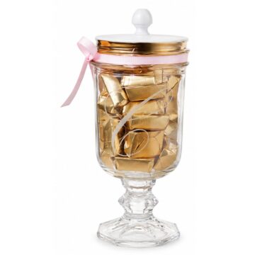 Glass pedestal candy jar filled with gold-wrapped chocolates, featuring an etched script monogram, white knob lid, and pink ribbon accent.