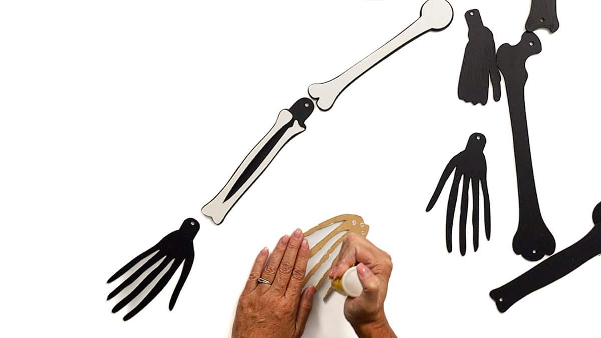 Hands gluing a laser cut skeleton hand piece while assembled black and white arm bones with joints are laid out on a white surface.