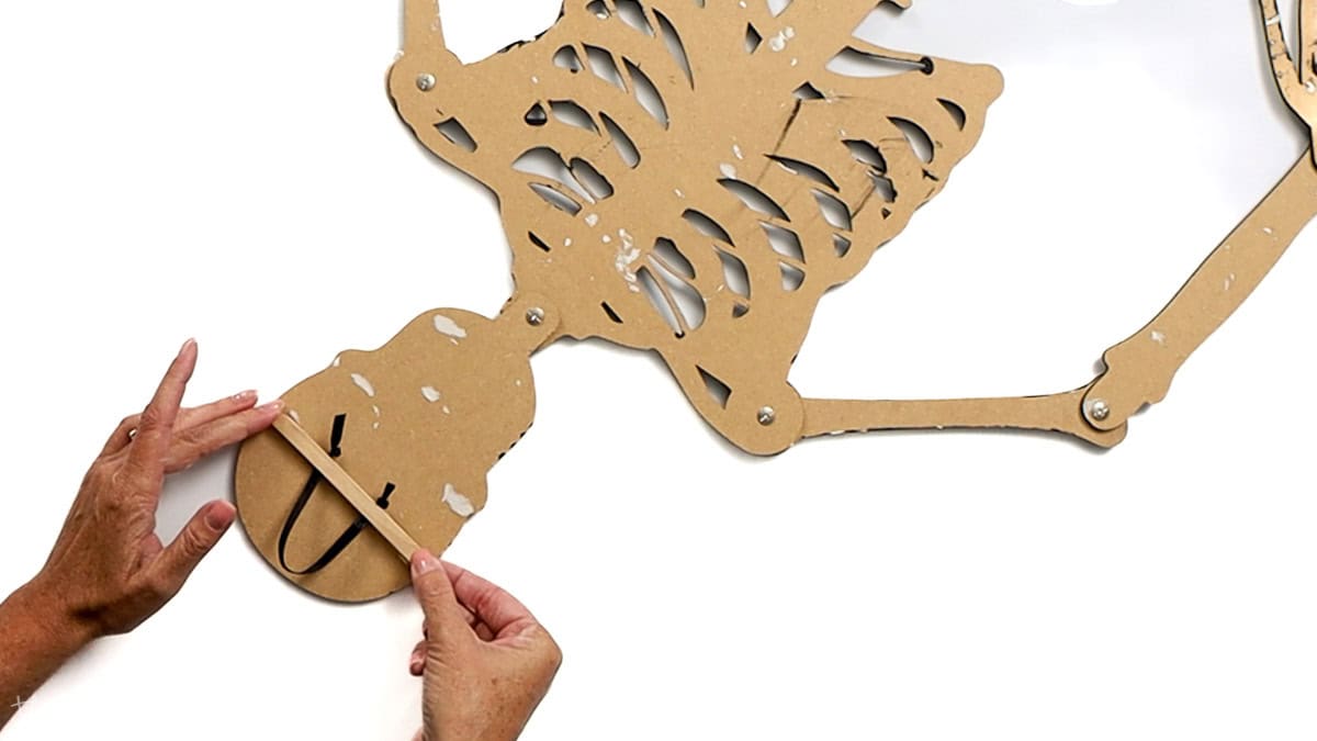Hands glue a wooden stick across the back of a paper skeleton's head to secure a black ribbon loop for hanging the decoration.