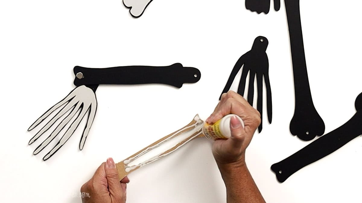 Hands applying glue to a laser cut skeleton forearm piece, with assembled black and white hand and arm bones visible on a white background.