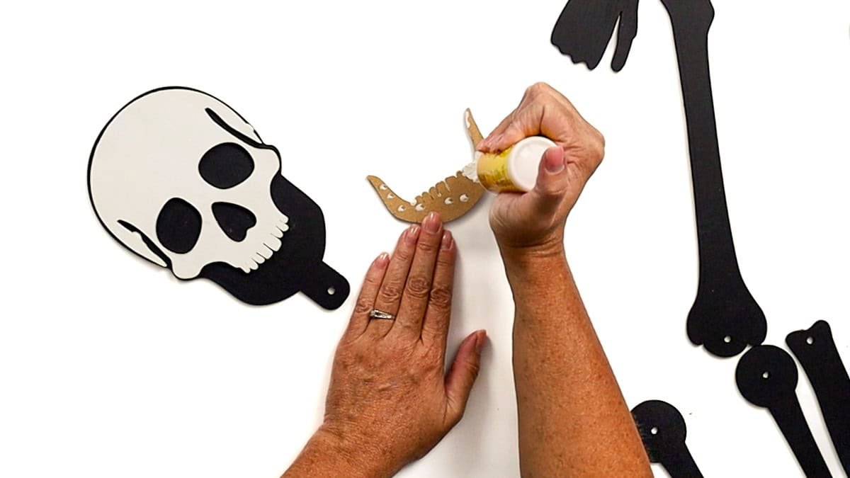 Hands applying glue to the back of a jawbone piece, with a painted black and white skull and other laser cut skeleton parts arranged on the table.