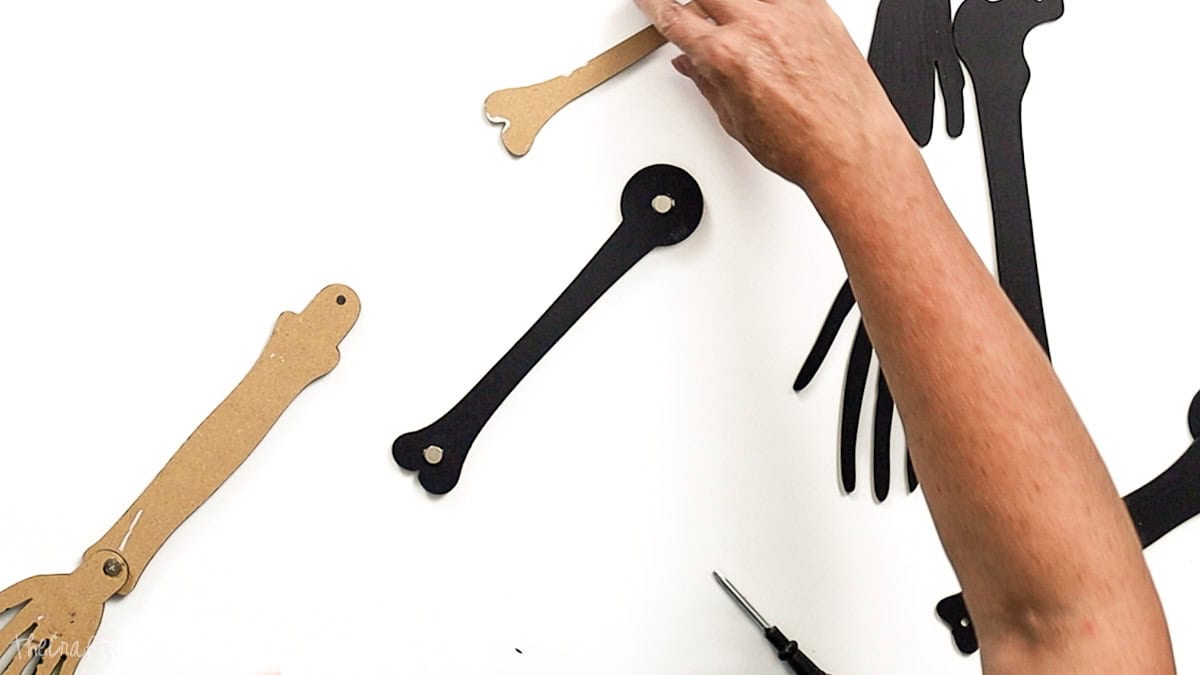 Hands assembling laser cut skeleton arm pieces, with screw casings connecting the black painted bones to brown backing pieces on a white surface.