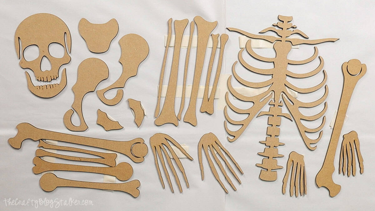 Laser cut MDF skeleton parts, including skull, ribs, pelvis, arms, legs, hands, and feet, laid out and taped in place for assembly.