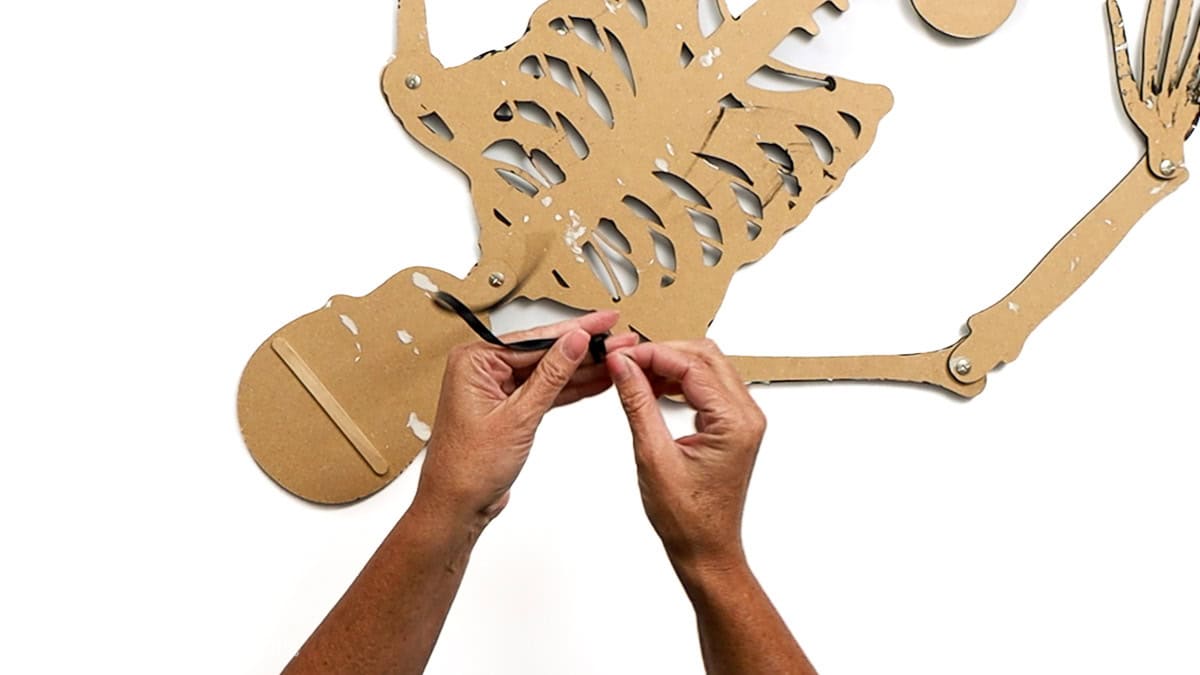 Hands tie a black ribbon into a knot at the back of a jointed paper skeleton craft, creating a hanger for displaying the decoration.
