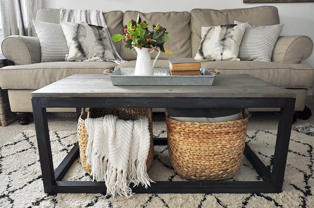 Modern farmhouse living room with a black-framed wooden coffee table holding baskets, books, and a tray with a white pitcher of greenery, in front of a beige couch.