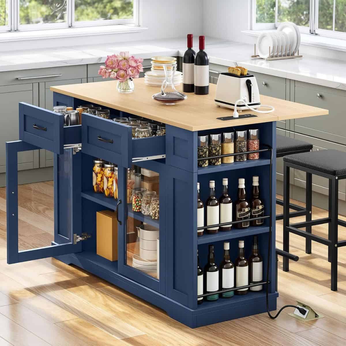 A blue kitchen island with wooden countertop, built-in storage shelves, drawers, and stools, styled with wine bottles, jars, and a toaster.