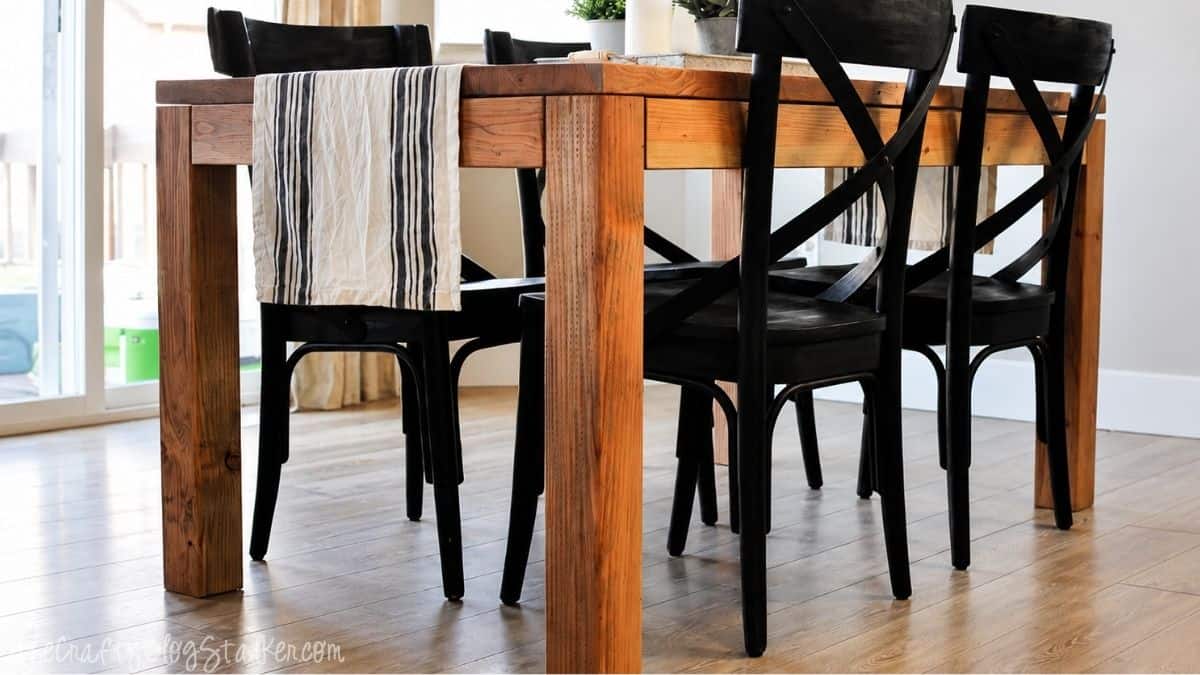 DIY wooden dining table with chunky legs and black chairs in a bright dining room with hardwood floors.