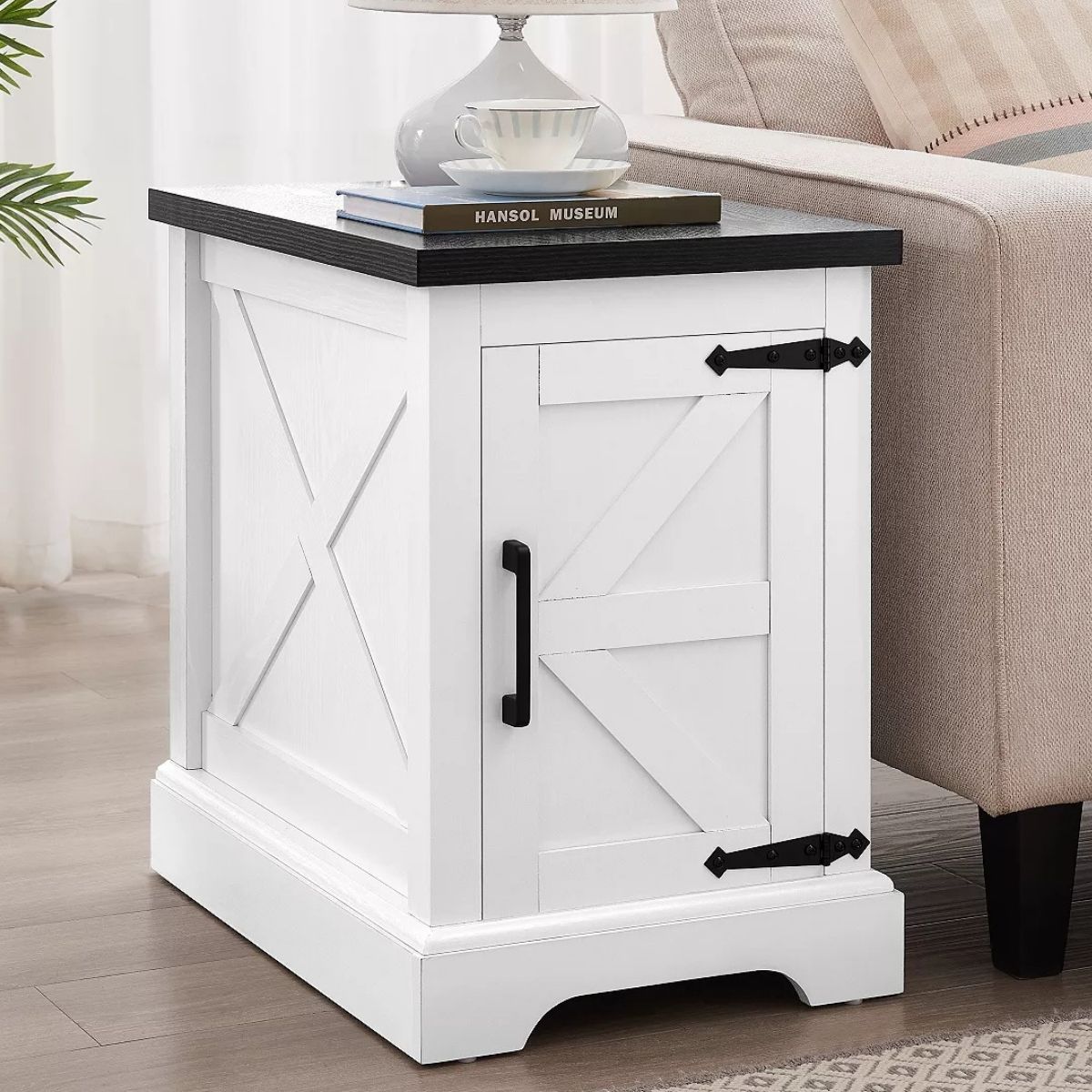 Farmhouse-style end table with a barn door cabinet, metal hardware, and a flat top holding books and a lamp, placed beside a sofa in a living room.