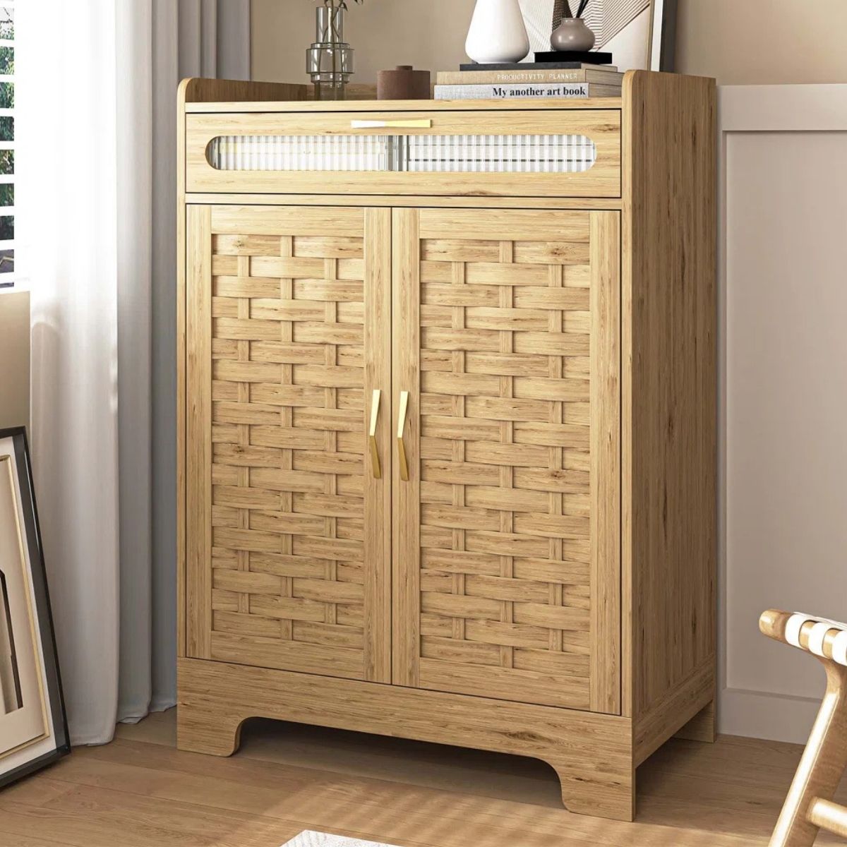 Wood storage cabinet with two woven panel doors, a top drawer, and decorative items on top, placed in a living room corner near a window.