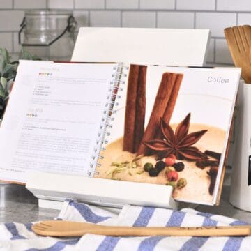 Open cookbook displayed on a white foldable recipe stand in a kitchen, surrounded by utensils, a striped towel, and farmhouse-style decor on a tiled backsplash.