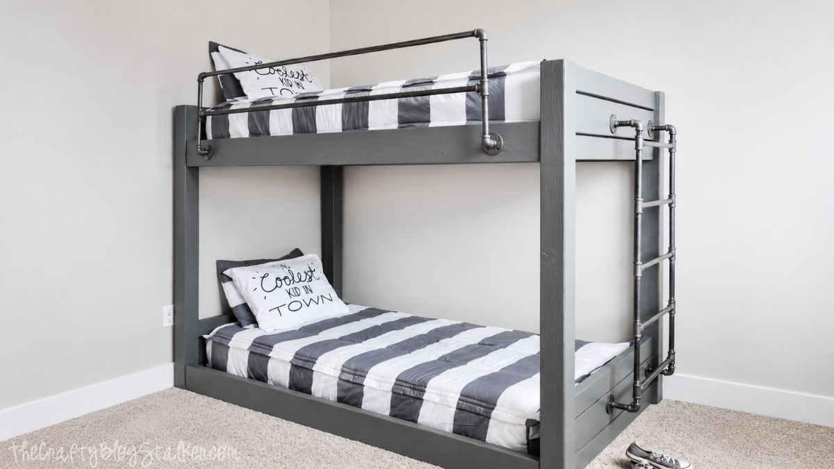 DIY industrial bunk bed with gray wood frame, metal pipe rails, ladder, and striped bedding in a modern bedroom.
