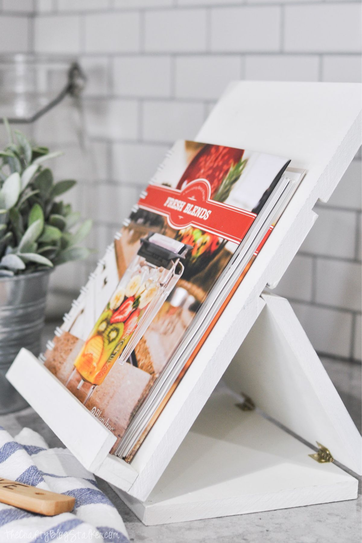 White wooden foldable recipe stand holding a cookbook titled "Fresh Blends," placed on a kitchen counter with subway tile backsplash and a small potted plant.