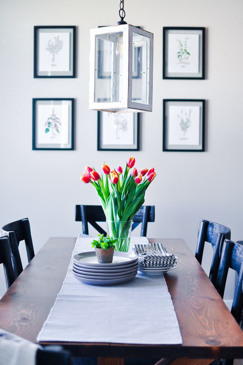 Dining table set with stacked plates, silverware, and a vase of red and yellow tulips, with framed botanical herb prints on the wall and a rustic lantern light fixture.